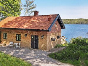 Une maison en briques avec un toit recouvert de tuiles rouges, une porte et deux fenêtres, située au bord d’un lac et dotée d’une terrasse en bois et de meubles d’extérieur.