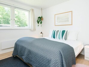 Et soveværelse med en seng, et vindue, en hængende plante, en sengetavlelampe og et billede i ramme på væggen.