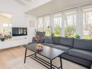 En moderne stue med en grå sofa, et sort sofabord, et fladskærms-tv og store vinduer.