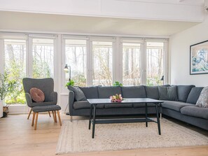 A modern living room with a grey sofa, a small table, and a chair. Large windows provide natural light and a view of trees.