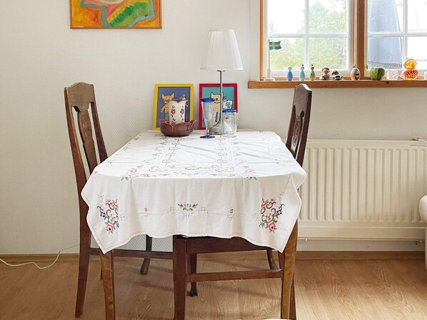 Une table à manger recouverte d’une nappe à motifs floraux, des chaises en bois et une fenêtre donnant sur des arbres.