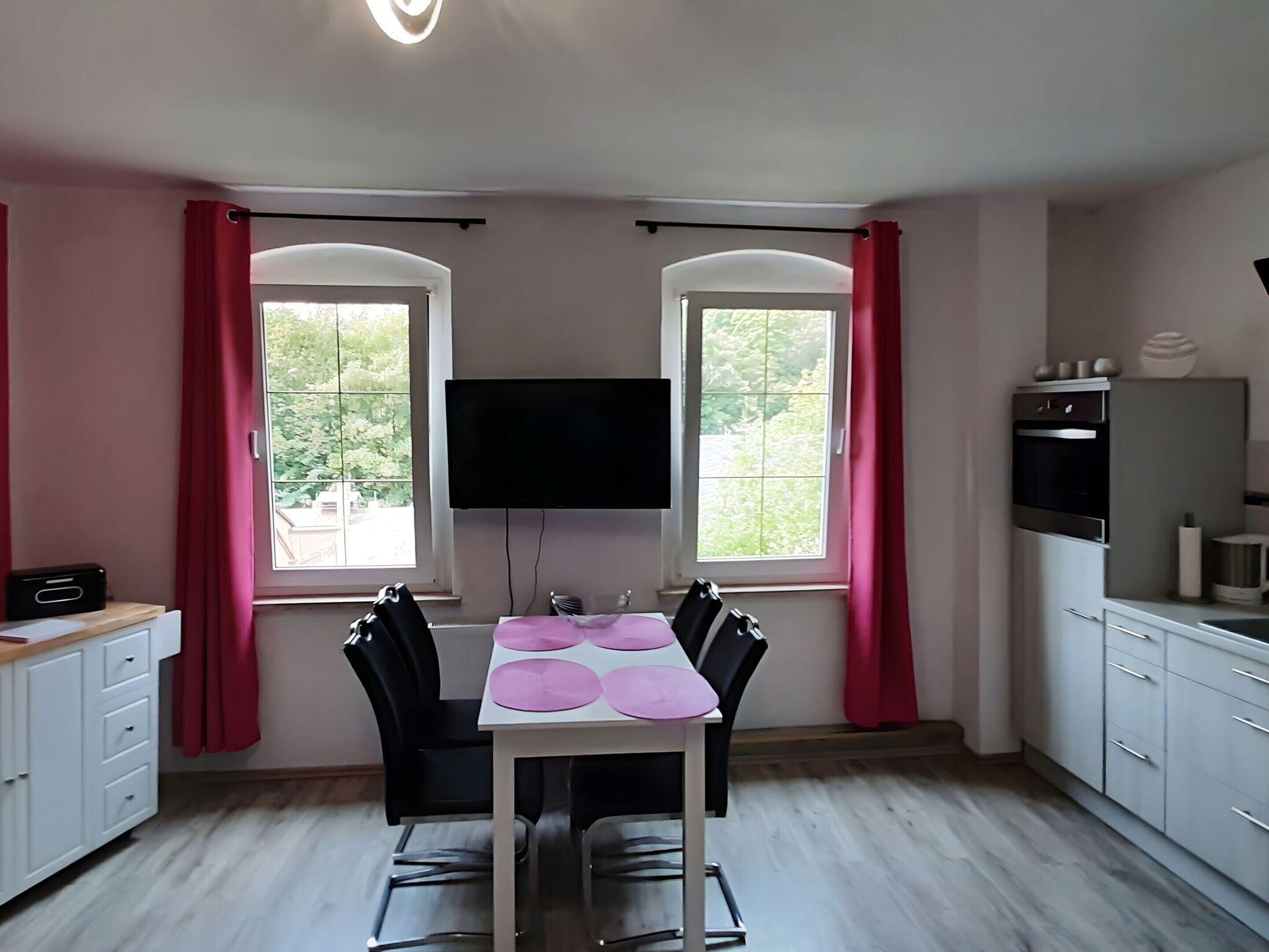 A compact kitchen with a dining table and chairs, a TV mounted above the table, and a window with curtains.