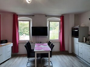 A compact kitchen with a dining table and chairs, a TV mounted above the table, and a window with curtains. - Lakeside Comfort With Garden (Schleiz)