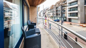 A balcony with wicker furniture, a view of the street, and modern buildings in the background. - Apartment With 2 Bedrooms and Large Terrace (Middelkerke)