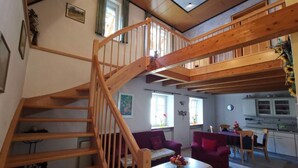 Un vaste espace de vie agrémenté d’un escalier en bois, d’une table à manger, de canapés violets et d’une cuisine à l’arrière-plan.