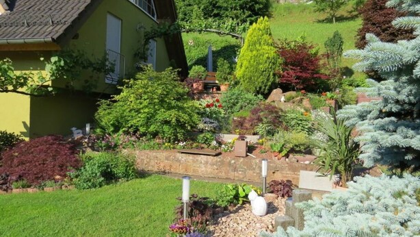 Une maison entourée d’un jardin bien entretenu, agrémenté de diverses plantes et d’un petit pont.