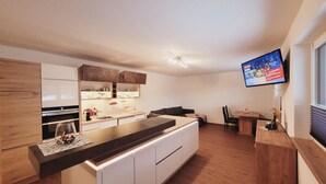 A modern kitchen with white cabinets, a wooden countertop, and a built-in oven. There is a dining area with a table and chairs, and a television mounted on the wall. - New Apartment in the Beautiful Pitztal (Sankt Leonhard im Pitztal)