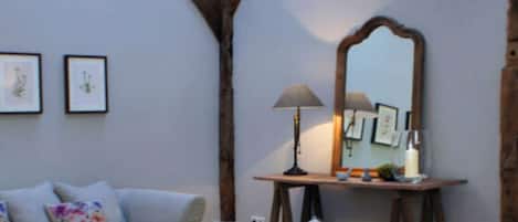 A cozy living room with exposed wooden beams, a wooden coffee table, a sofa with floral cushions, a side table with a lamp, and a mirror.