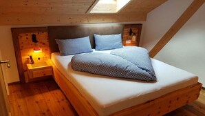 Un lit en bois, recouvert d’une courtepointe à carreaux, dans une pièce au plancher de bois et au plafond en pente.