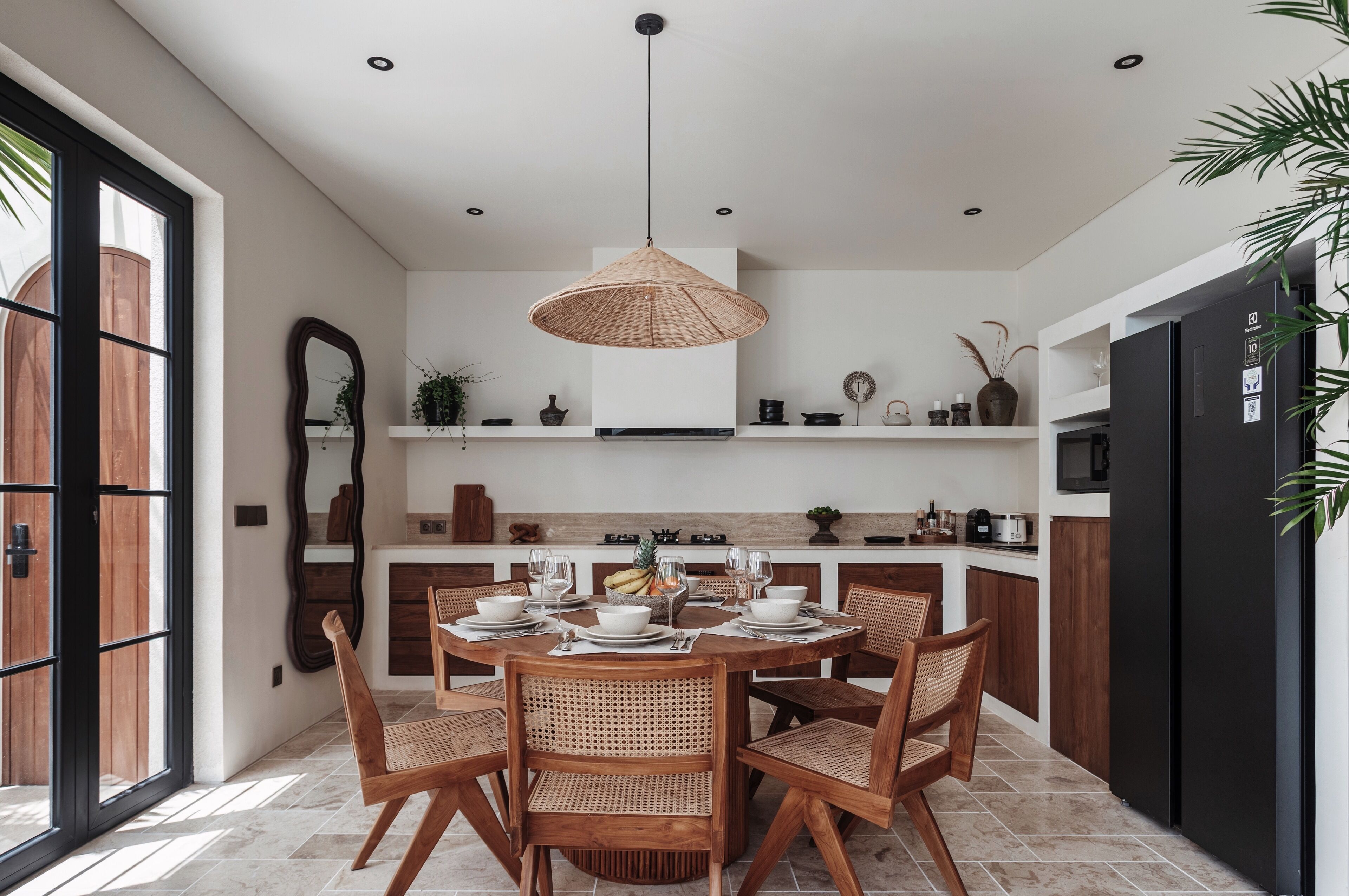 A modern kitchen with a dining area, featuring a round table, wicker chairs, and a hanging light fixture.