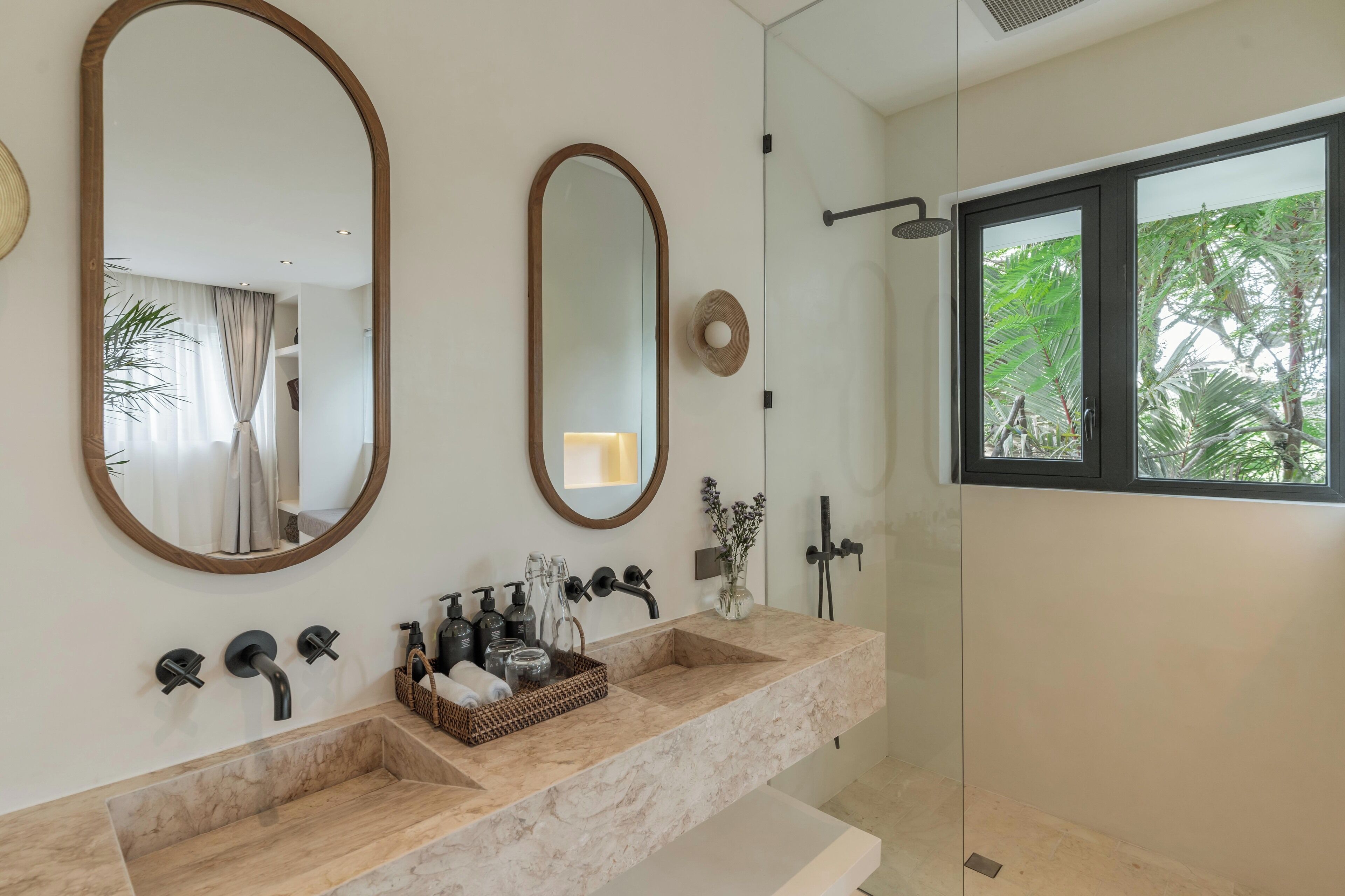 A modern bathroom with a marble vanity, two large mirrors, a glass shower enclosure, and a view of greenery outside.