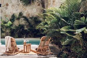 A poolside area with wicker chairs, a small table with a bottle and glasses, and lush tropical plants. - The Revery Bali (Canggu)