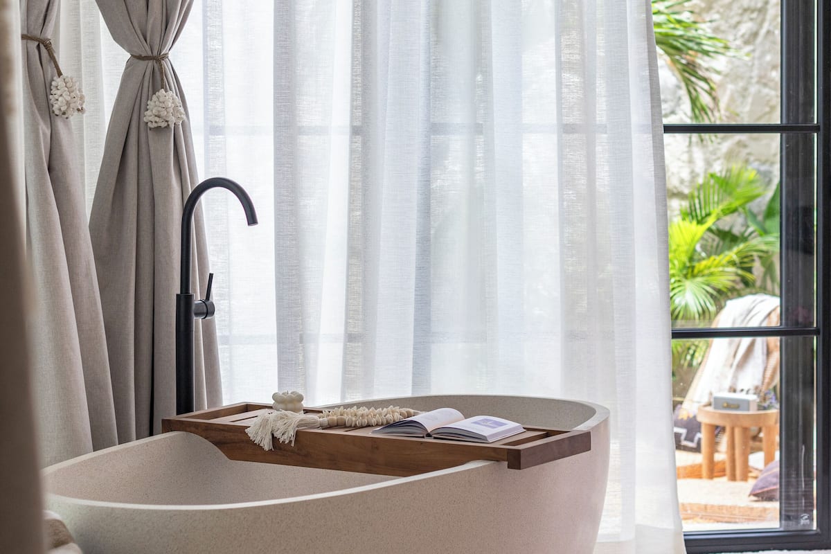 A modern bathroom with a freestanding bathtub, a wooden side table, and a large window with sheer curtains.