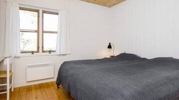 A bedroom with a wooden floor, a bed with a grey blanket, a window with white curtains, and a bedside table with a lamp.