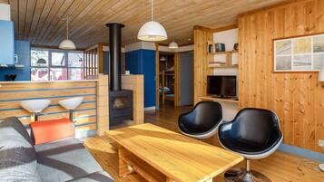 A modern interior with a wooden ceiling, a central fireplace, a sofa, two black chairs, a wooden table, and a kitchen area with a window.