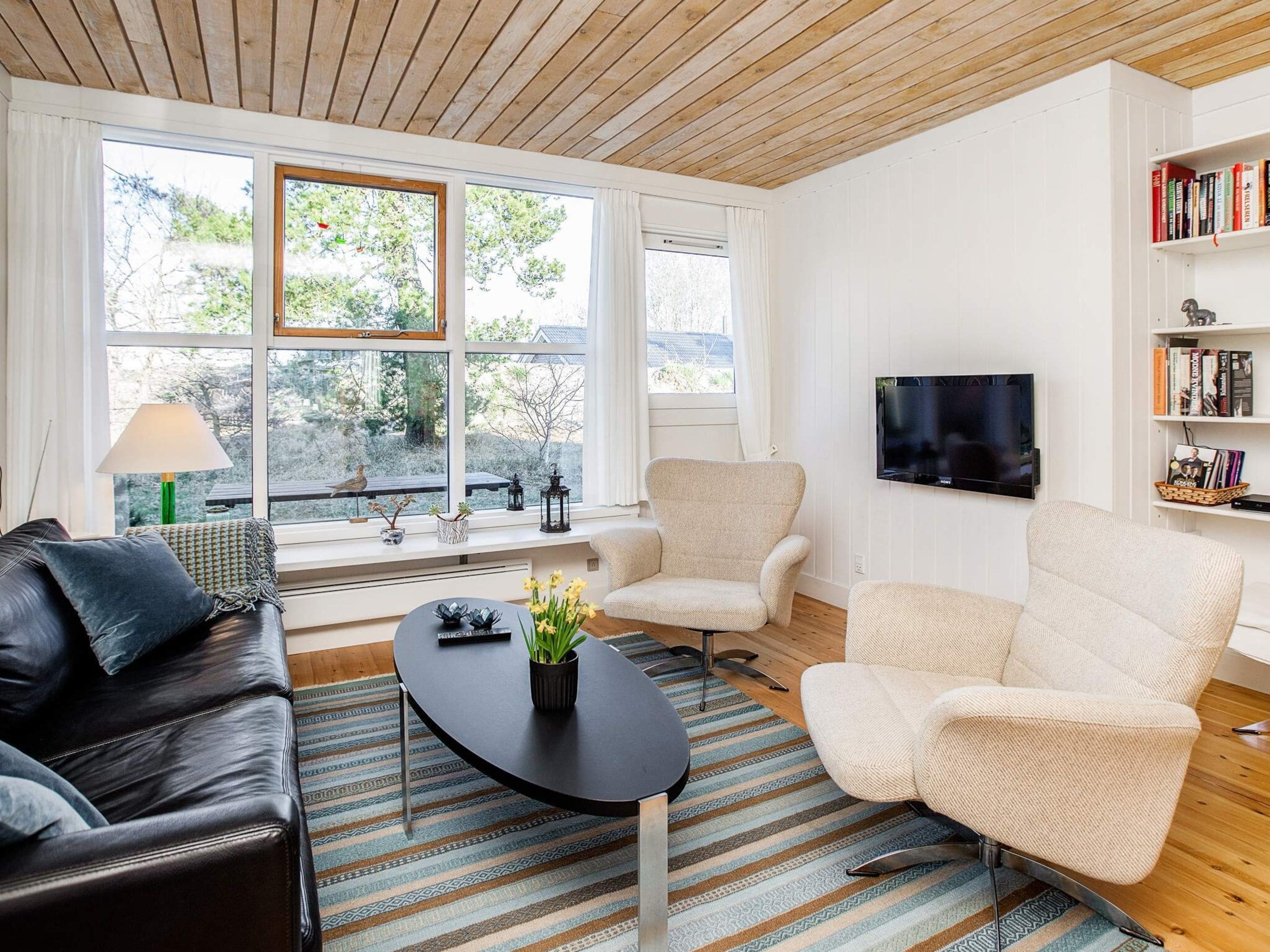 Una sala de estar moderna con un sofá de cuero, dos sillones, una mesa de centro y un televisor montado en la pared. Amplios ventanales ofrecen luz natural y vistas al exterior.
