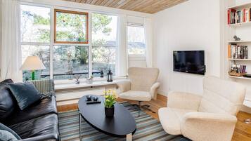 Una sala de estar moderna con un sofá de cuero, dos sillones, una mesa de centro y un televisor montado en la pared. Amplios ventanales ofrecen luz natural y vistas al exterior.