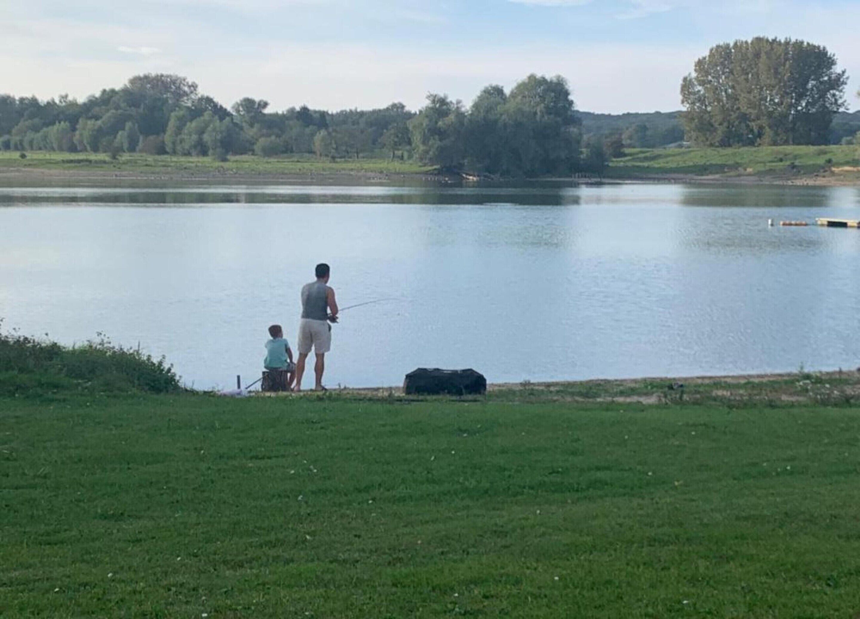 Un lago tranquilo con orilla cubierta de hierba, un hombre pescando y un niño cerca.