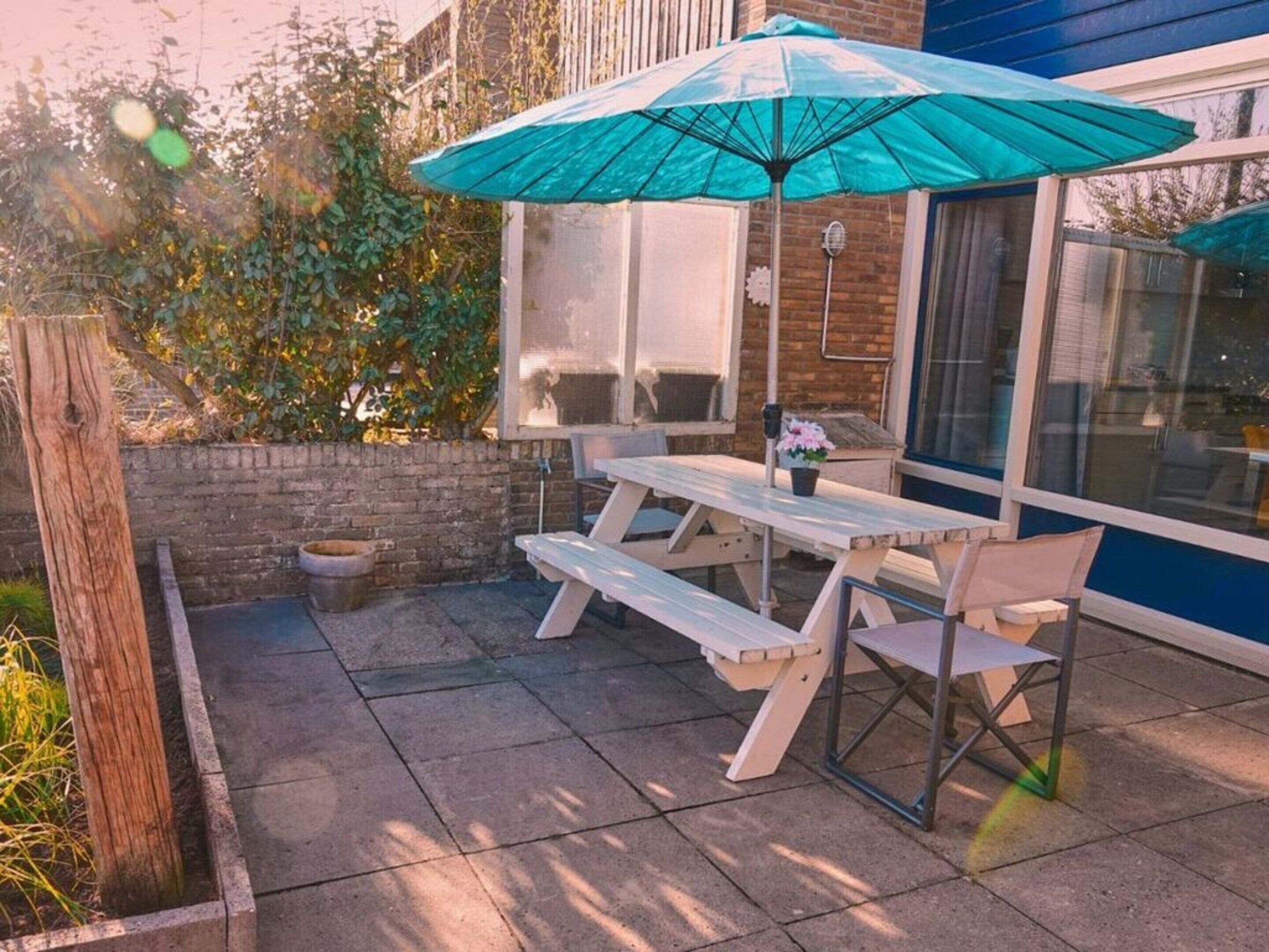 En udendørs terrasse med et picnicbord og stole, en blå parasol samt en murstensmur.