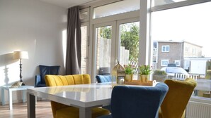 A modern dining area with a white table and yellow and blue chairs, large windows with curtains, and a view of the street outside.