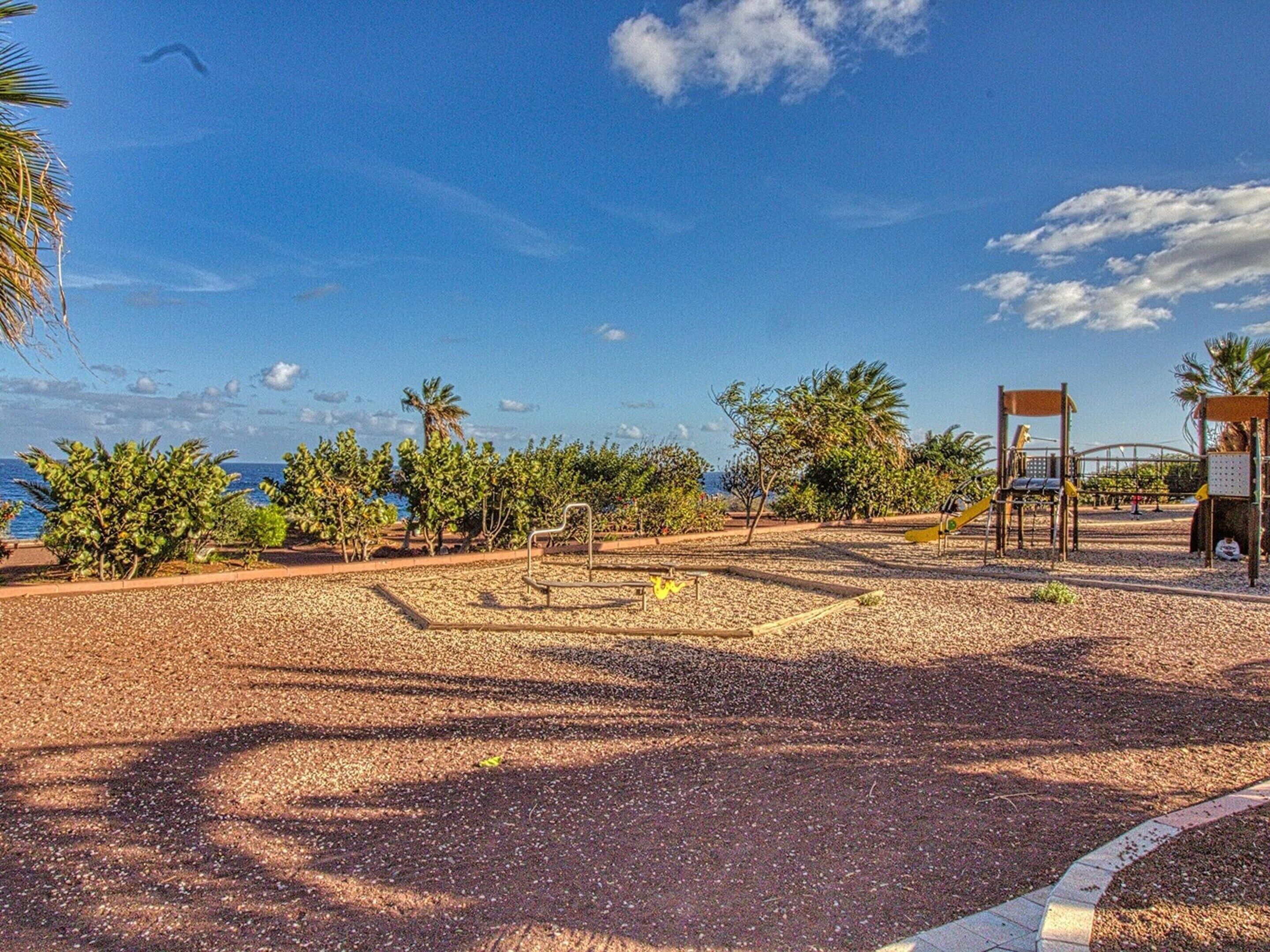 Un patio de juegos al aire libre con estructuras para escalar y una zona de estar, rodeado de árboles y un cielo despejado.