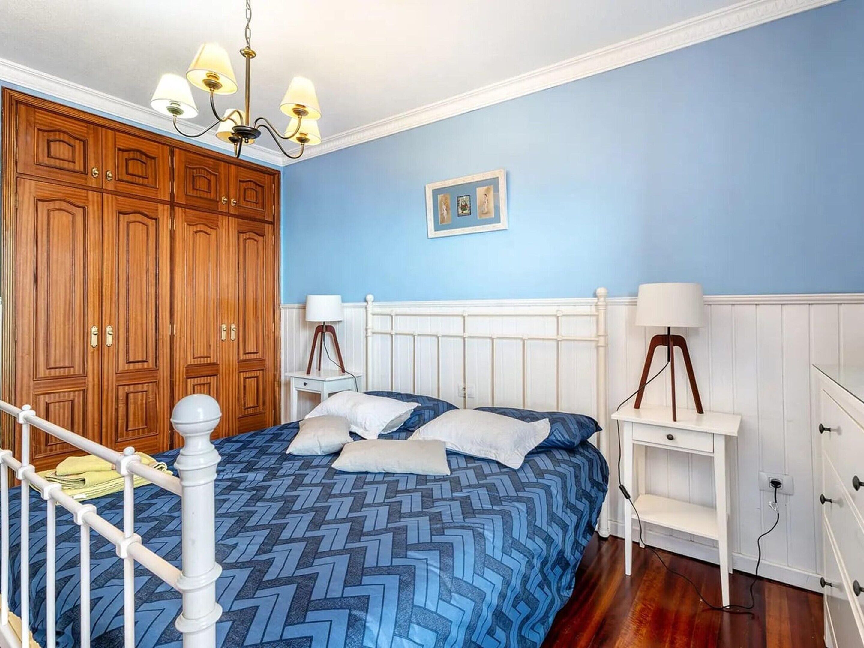 A bedroom with a bed, wooden wardrobe, white bedside tables, and a chandelier.