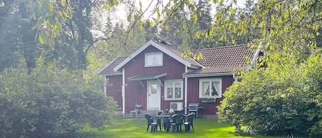 Une maison rouge avec une porte et des fenêtres blanches, entourée d’herbe verte et d’arbres.