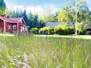 Une maison rouge avec un toit en pente, entourée d’un champ d’herbes hautes et bordée d’arbres au loin.