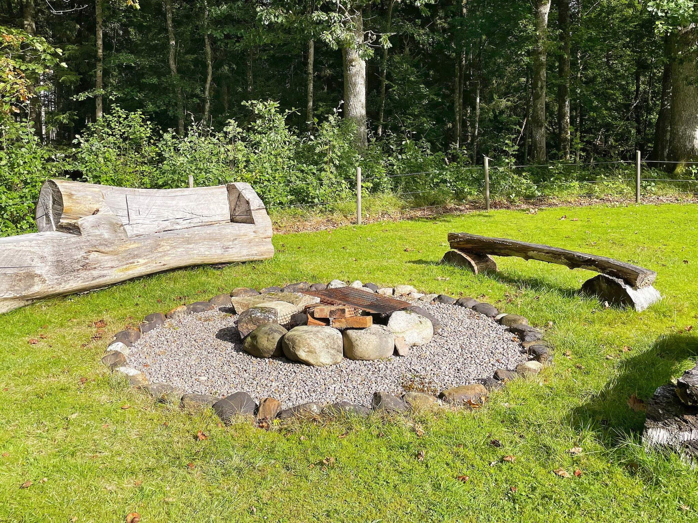 Un foyer circulaire en pierre, entouré d’une zone herbeuse avec des bûches et un banc en bois.