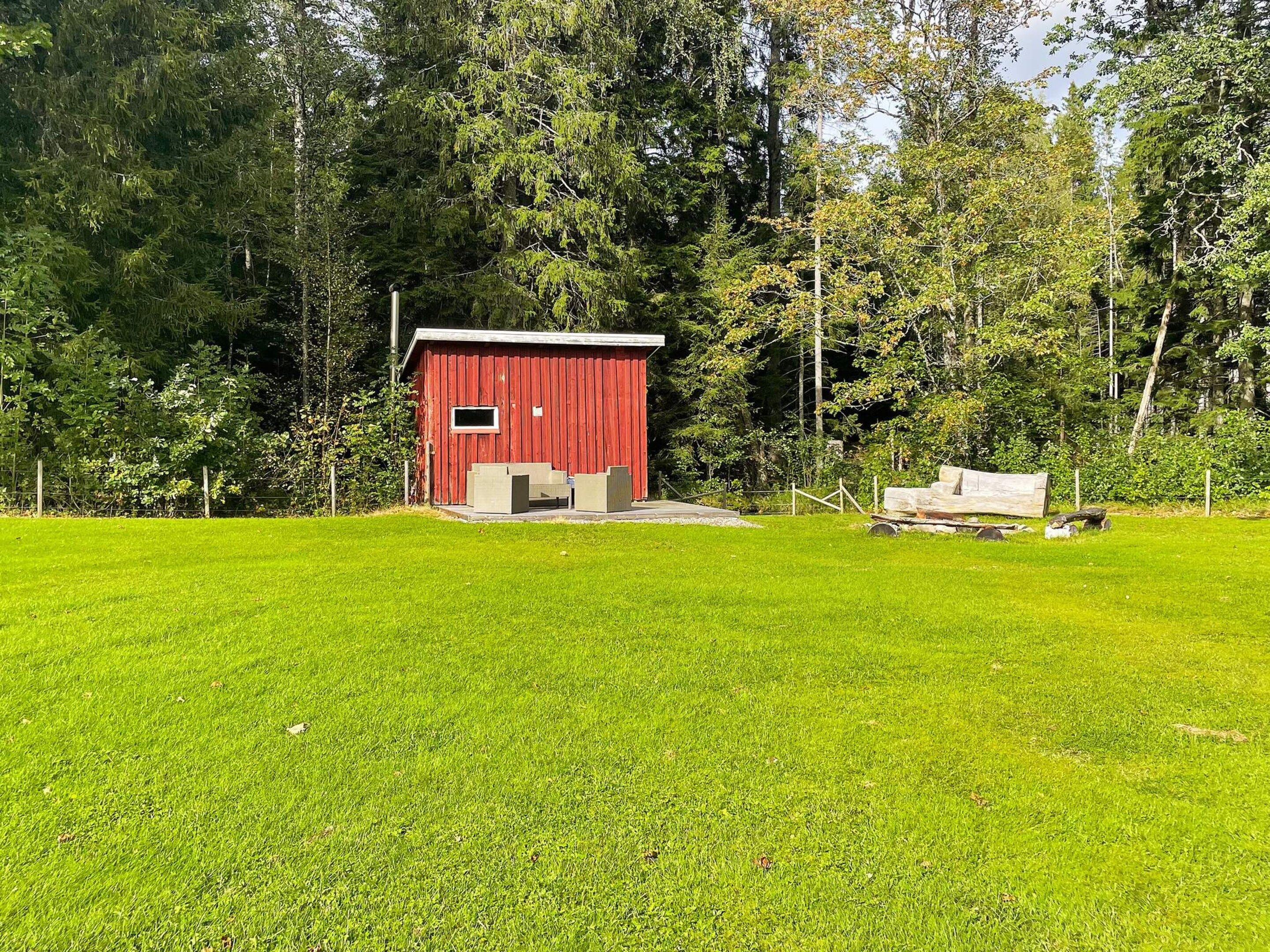 A red wooden structure with a small window, surrounded by a well-maintained grassy area and dense trees.