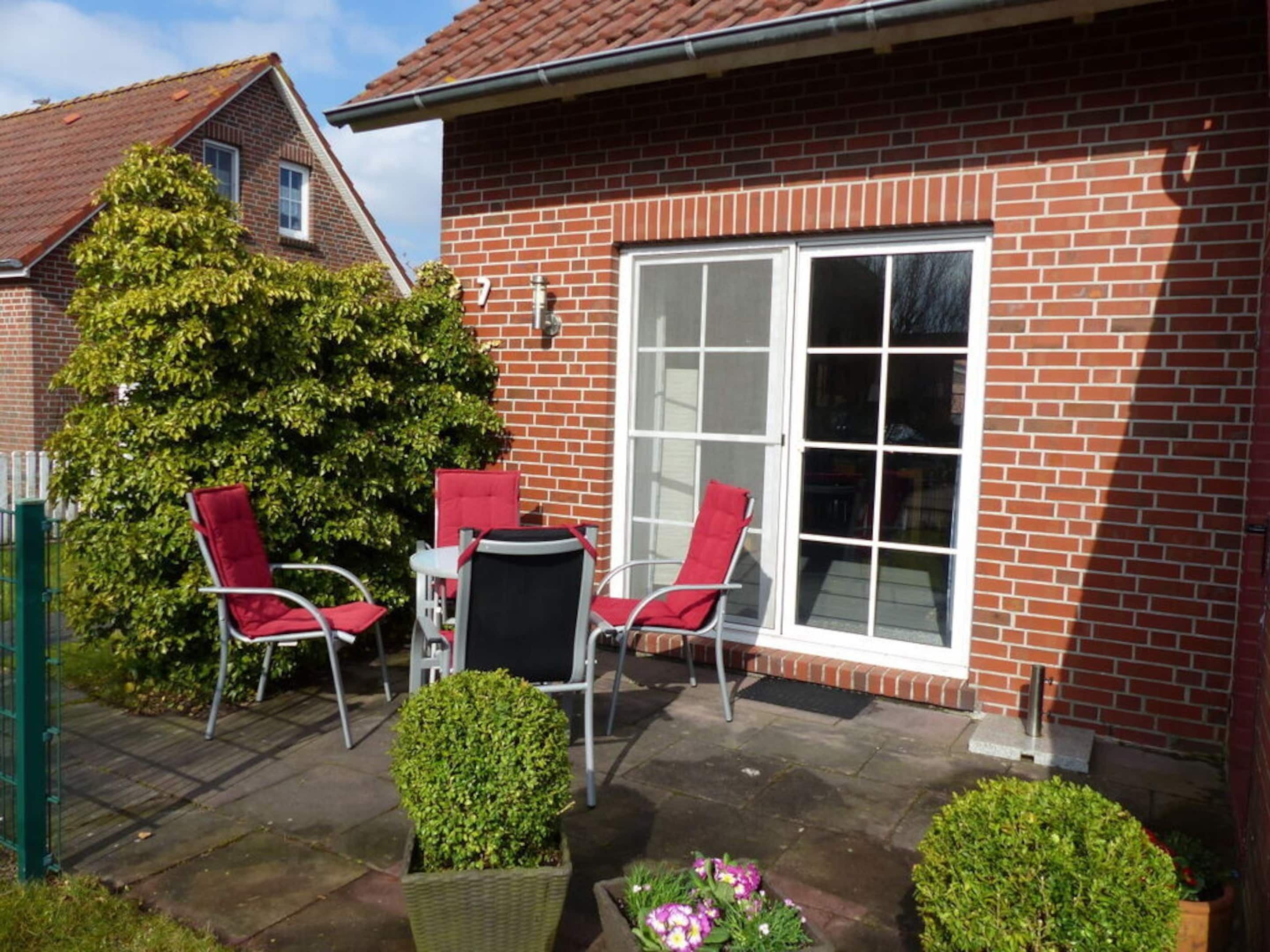 A brick house with a large window, a patio with two red chairs and a table, and a small garden with a hedge and flowers.