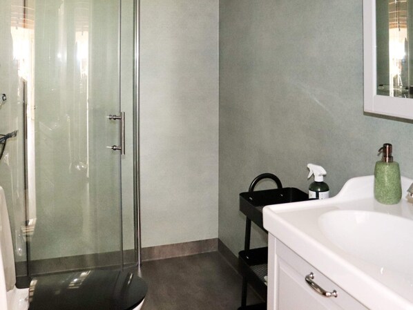 Une salle de bain avec une douche à parois de verre, un lavabo blanc doté d’un distributeur de savon vert et un porte-serviettes noir.