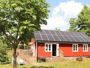 Un bâtiment rouge surmonté d’un toit équipé de panneaux solaires, avec une terrasse en bois et un arbre au premier plan.
