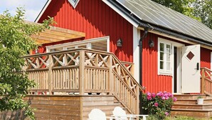 Een rood gebouw met een houten terras, een witte deur en een kleine tuin met roze bloemen.