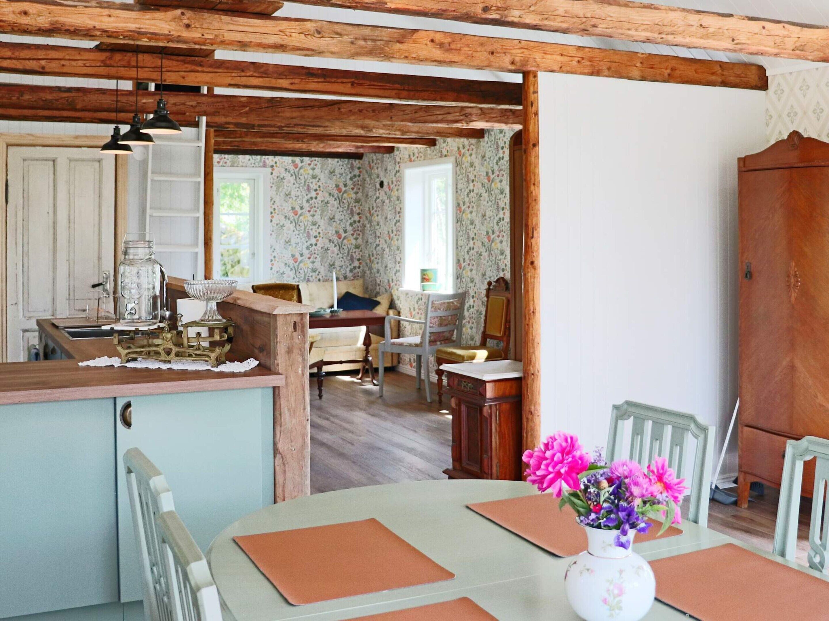 A rustic kitchen with wooden beams, a dining table with chairs, and a floral wallpapered room in the background.