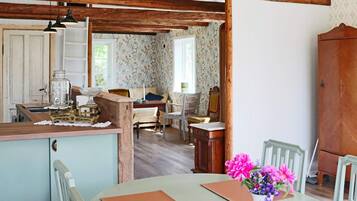 A rustic kitchen with wooden beams, a dining table with chairs, and a floral wallpapered room in the background.