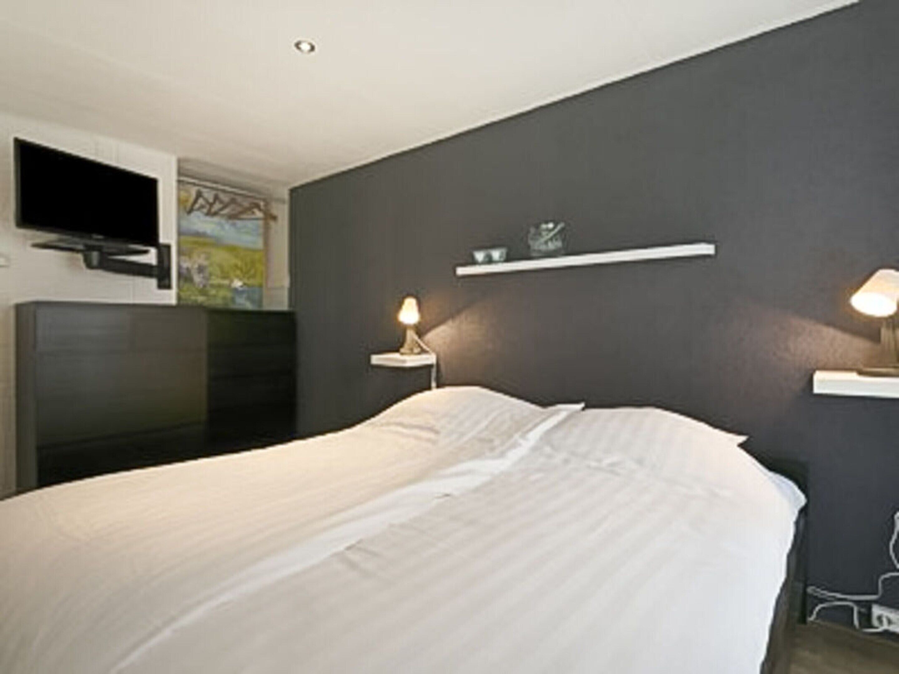 Un dormitorio con cama, televisor montado en la pared, mesita de noche con lámpara y estante sobre la cama.