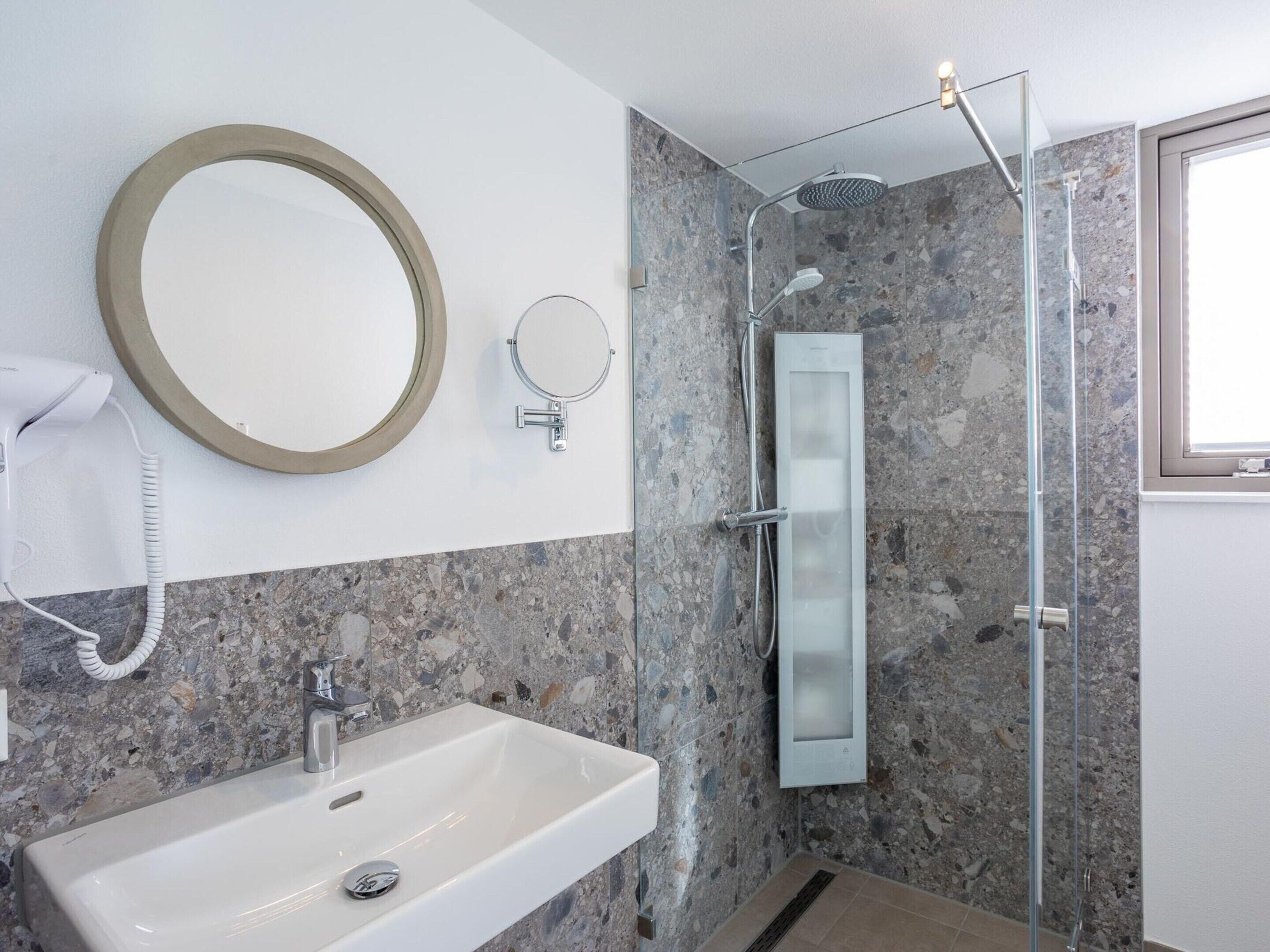 Une salle de bain moderne avec un lavabo rectangulaire, un miroir rond, une douchette à main et une douche à parois de verre.