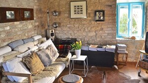 A rustic room with a stone wall, a sofa with patterned cushions, a small table with a vase of flowers, and a window with a view of greenery. - Lovely Apartment in Ripatransone (Ripatransone)