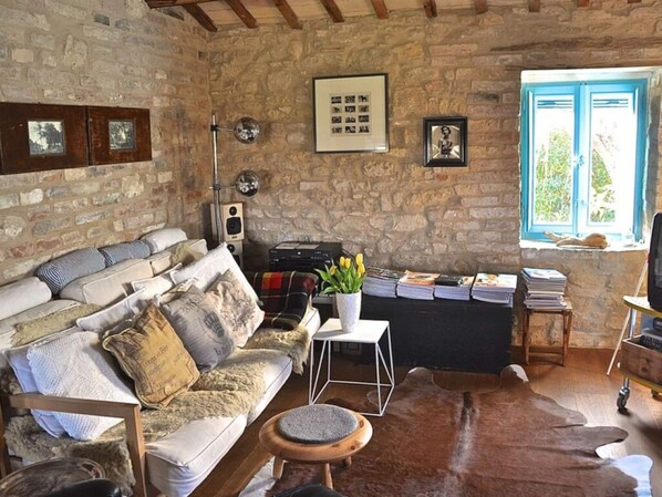 A rustic room with a stone wall, a sofa with patterned cushions, a small table with a vase of flowers, and a window with a view of greenery.