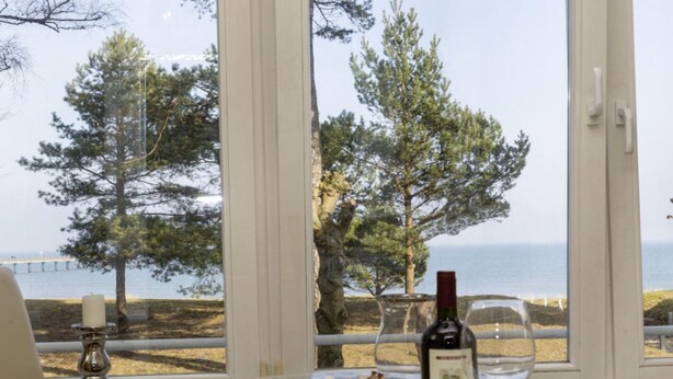A table with a plate of food, a bottle of wine, and a glass, set against a backdrop of a scenic view through large windows. - Sea Lymphony - in Villa Frigga (Binz)