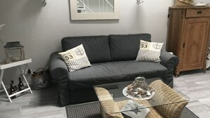 A living room with a grey sofa, two cushions, a wicker coffee table, a small side table, a cabinet, and a framed picture on the wall. - Holiday Apartment Inselstern (Fehmarn)