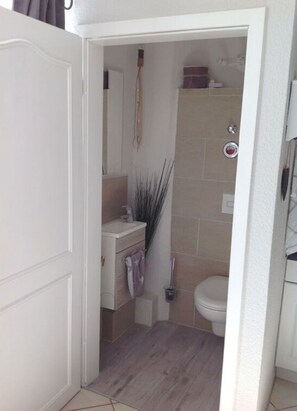 A narrow bathroom with a white door, a wooden vanity, a toilet, and a shower area. - Holiday Apartment Inselstern (Fehmarn)