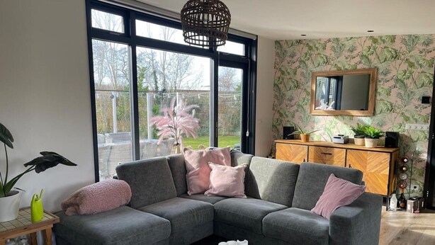 Ein modernes Wohnzimmer mit einem grauen Ecksofa, einem Couchtisch aus Holz mit Kerzen und einem großen Fenster mit Blick ins Freie.