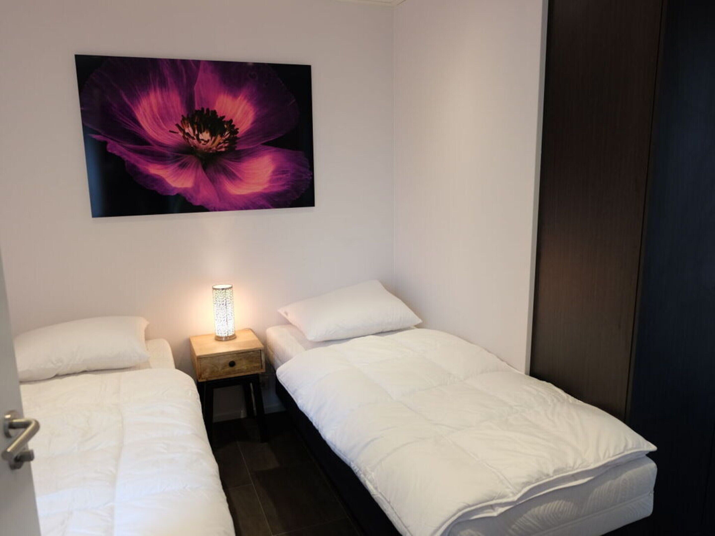 A small, neatly arranged bedroom with two beds, a bedside table with a lamp, and a large flower painting on the wall.