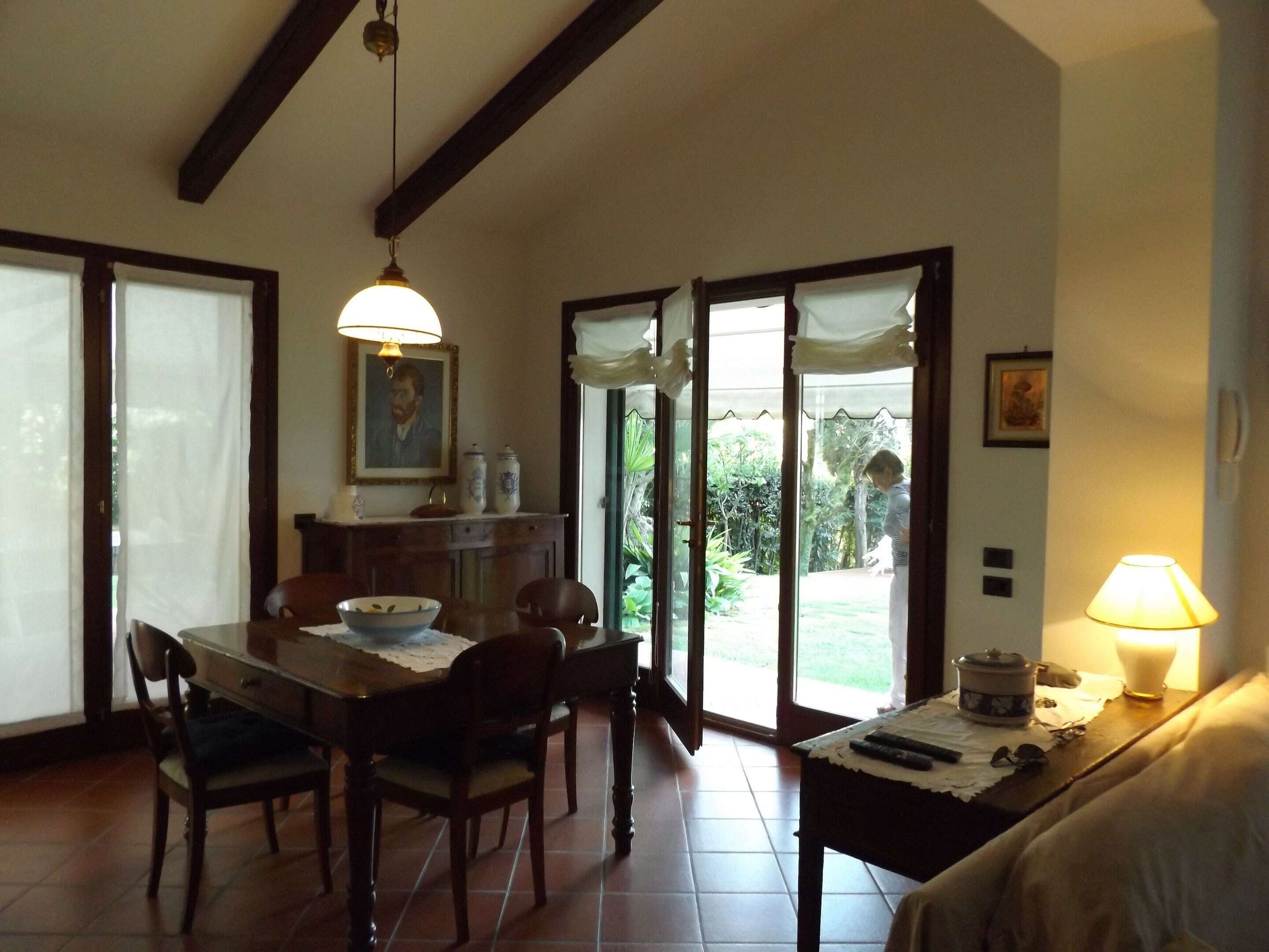 A dining area with a wooden table, chairs, and a lamp. There is a window with curtains and a view of greenery outside.