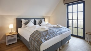 A neatly made bed with white linens and plaid bedding, a bedside table with a lamp, and a large window with a view of the outdoors. - Thatched Villa With Sauna and Fireplace (Dranske)