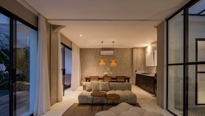 Uma sala de estar moderna com sofá, banquinho e duas mesinhas laterais. Há uma área de jantar com mesa e cadeiras de madeira, e uma cozinha com armários e bancada.