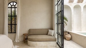 Une pièce d’angle avec un banc rembourré, un petit tabouret, une grande fenêtre avec des rideaux et une vue sur un mur blanc orné d’arches et de vases.