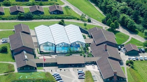Eine Vogelperspektive zeigt einen Gebäudekomplex mit einem zentralen Schwimmbecken, umgeben von mehreren kleineren Gebäuden, einem Parkplatz und grünen Rasenflächen.